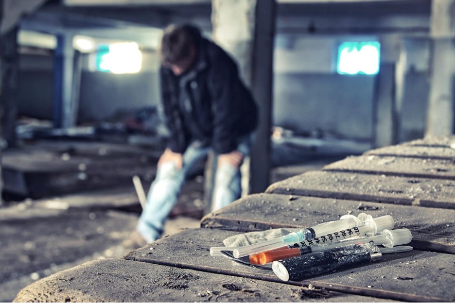 Unhoused individual in the background with heroin, needle, and lighter in the foreground